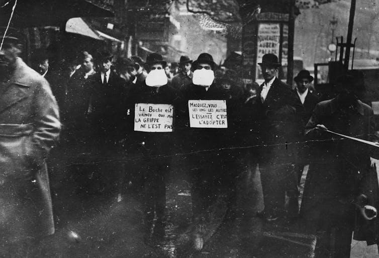 Two men wearing and advocating the use of flu masks in Paris with a crowd of people behind them.