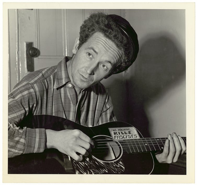 A man with a hat playing a guitar with a sticker attached that says, 'This Machine Kills Fascists.'