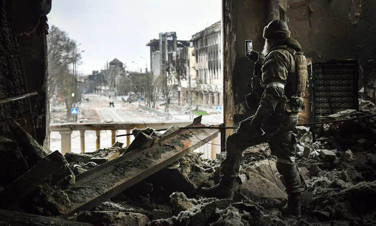 A person stands on rubble and holds up a phone, facing toward an opening in the wall and a street.