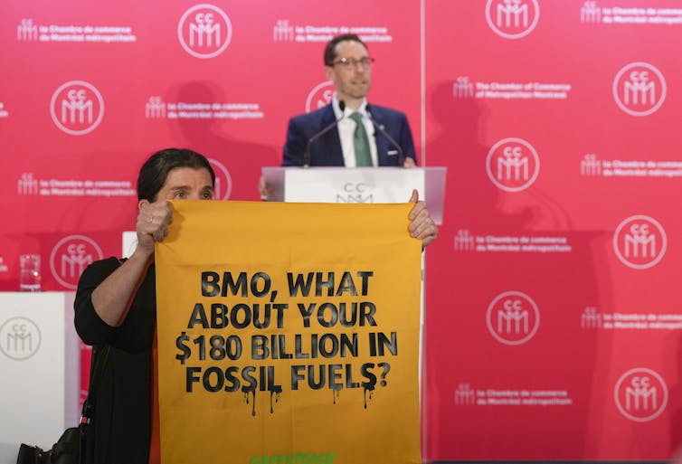 A man carries an orange sign that reads BMC what about your $100 billion in fossil fuels in front of a man standing behind a podium.