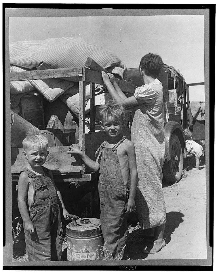 A family on the road, standing next to a rickety truck with their belongings. Two boys in overalls are wearing no shirts.