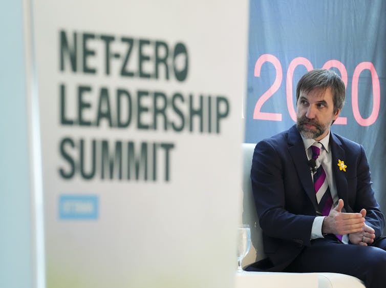 A bearded man in a suit sits next to a sign that reads Net-Zero Leadership Summit.