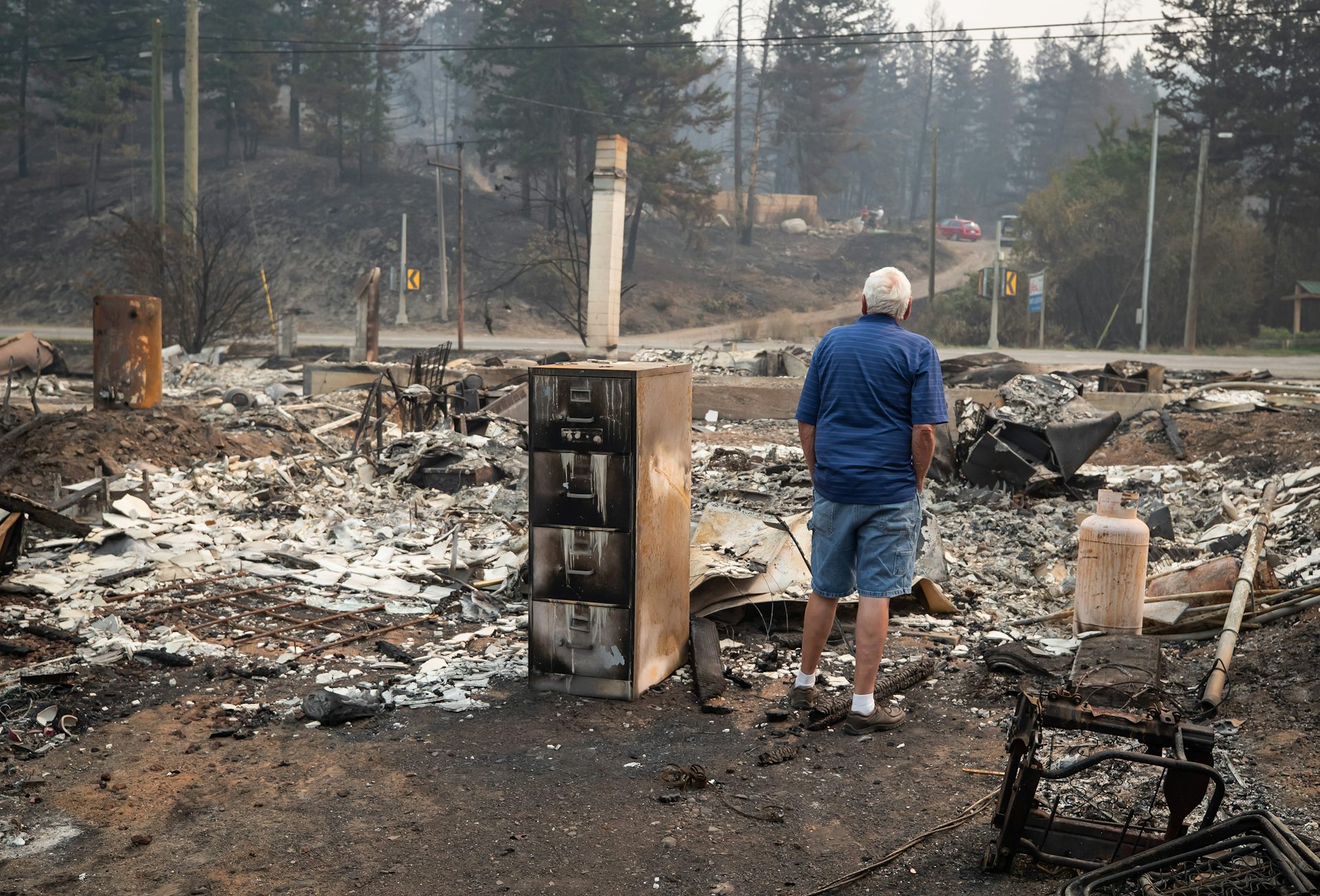 A grey-haired man is seen from behind as he looks at the burnt remains of a house.