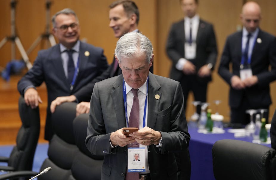 a white guy with gray hair and a gray suit named jerome powell looks down at his phohne in front of several other white men in suits