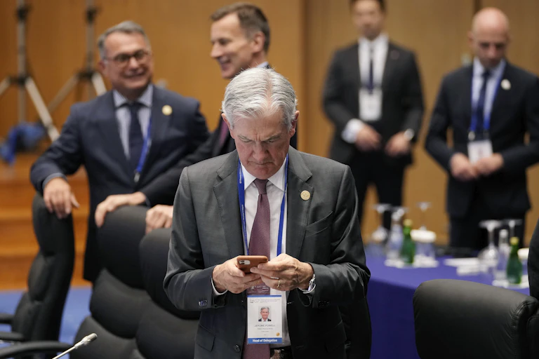 a white guy with gray hair and a gray suit named jerome powell looks down at his phohne in front of several other white men in suits