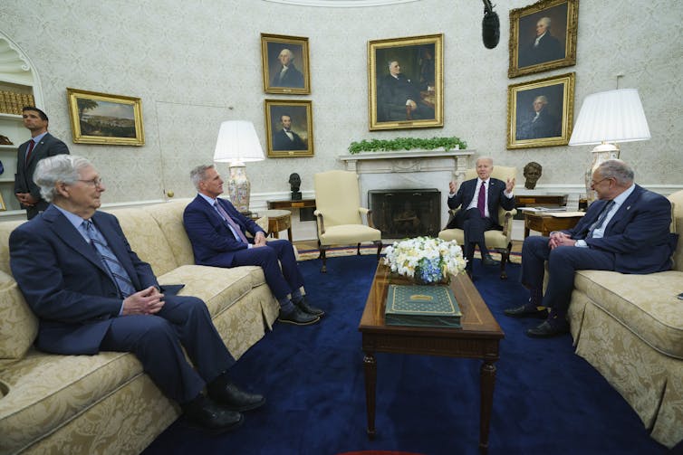 Four white men sit on white couches in a large office filled with presidential portraits.