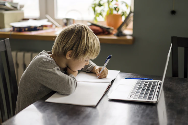 Sitting at a desk, and with his laptop in front of him, a young boy writes in his notebook.