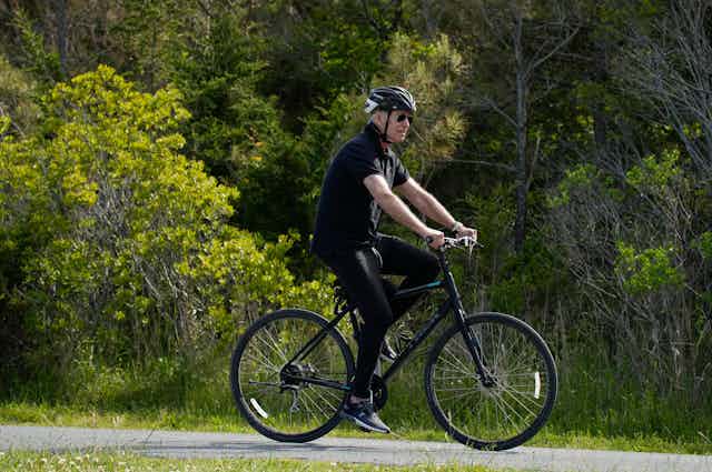 An aged man rides a bike along a tree-lined path. He is dressed in a black T-shirt and pantsand is wearing a black bike helmet.