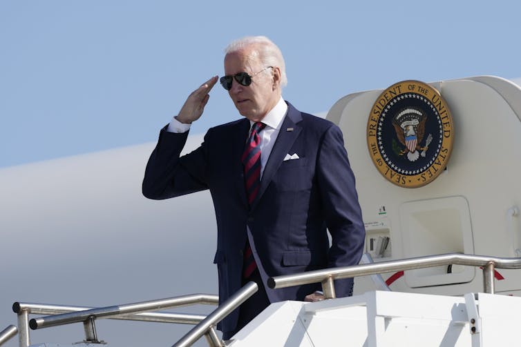 A grey-haired man in a dark suit and sunglasses salutes as he disembarks a plane.