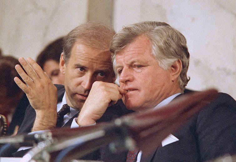 A balding man speaks into the ear of another man with a full head of wavy salt-and-pepper hair.