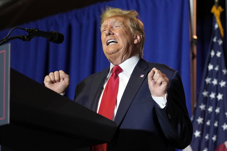 A blond man in a blue suit and red tie grimaces as he gestures in front of a podium.