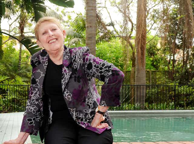 a smiling short-haired woman by a swimming pool, wearing a purple and black patterned jacket, and jeans
