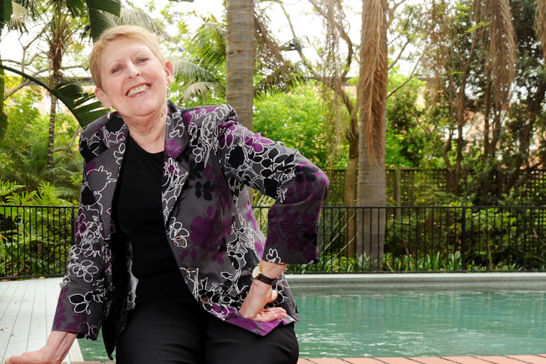 a smiling short-haired woman by a swimming pool, wearing a purple and black patterned jacket, and jeans