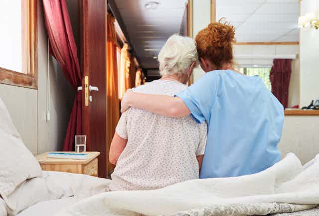A woman with red hair in medical scrubs seen from behind, with her arm around a woman with gray hair sitting on a bed