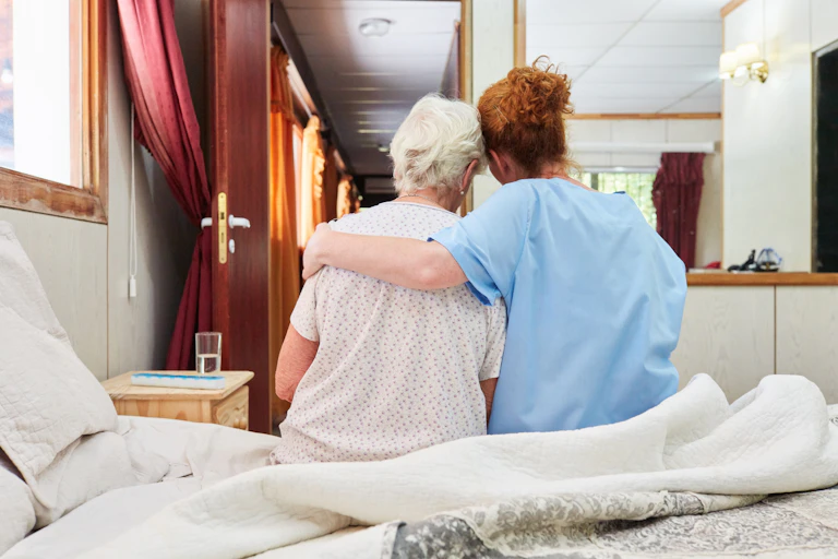 A woman with red hair in medical scrubs seen from behind, with her arm around a woman with gray hair sitting on a bed