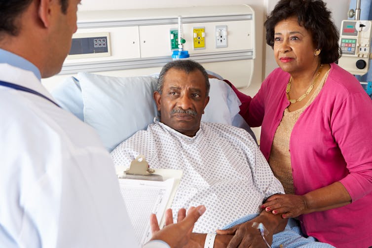 A man in a hospital bed and a woman with her arm around him, both looking at a man in a white coat seen from behind