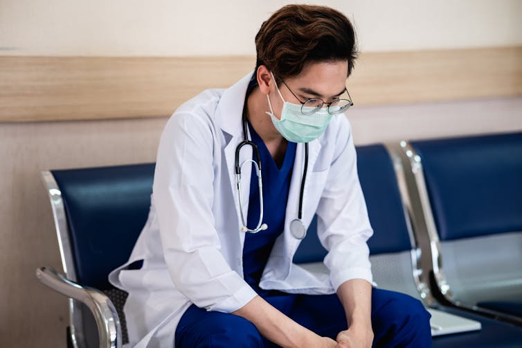 A man in a white coat, stethoscope and face mask sitting in a chair and looking upset