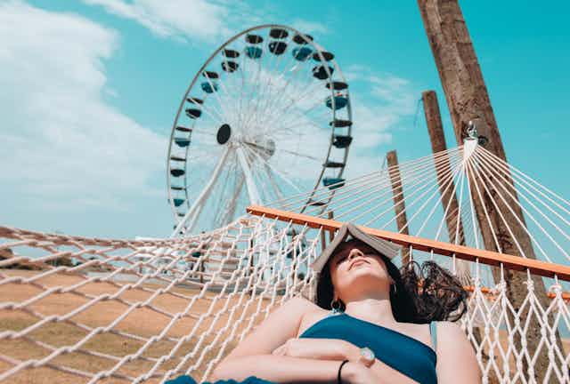 Woman lies dozing in hammock with book over face and ferris wheel in background