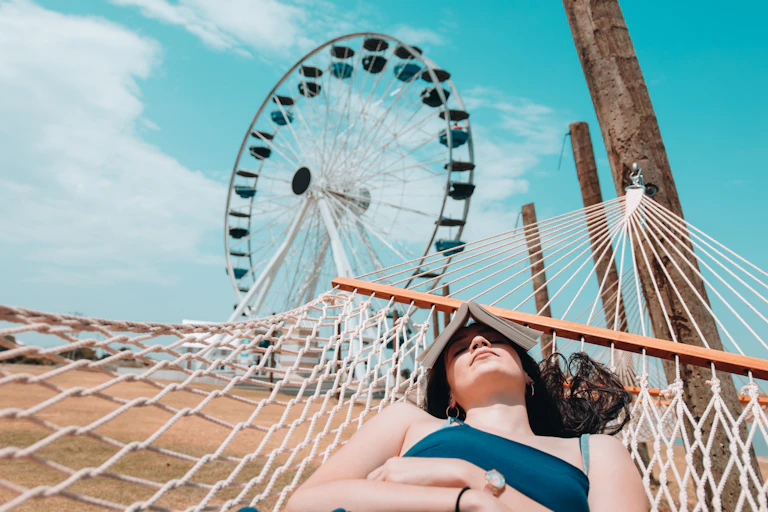 Woman lies dozing in hammock with book over face and ferris wheel in background