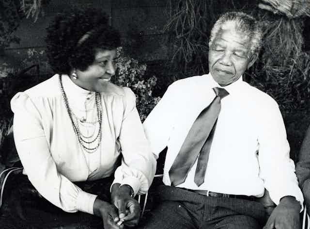 A black and white photo of a couple sitting outdoors in a garden and holding hands, smartly dressed. She smiles while he looks bemused.