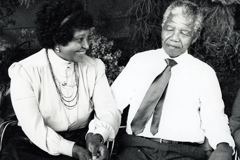 A black and white photo of a couple sitting outdoors in a garden and holding hands, smartly dressed. She smiles while he looks bemused.