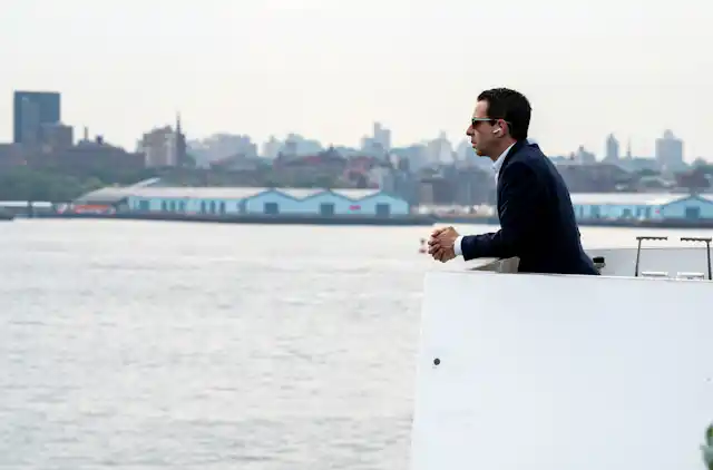Kendall Roy wearing AirPods on a boat, in a black suit.