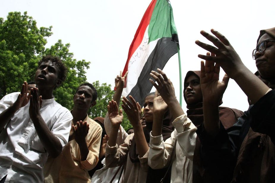 A group of five people clapping their hands or making "peace" signs, congregated around a Sudanese flag