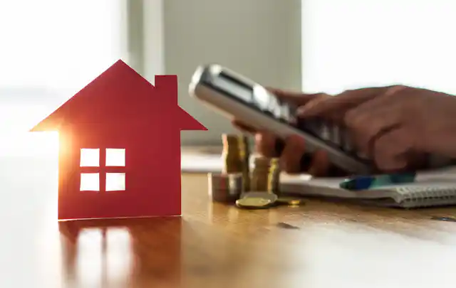 Background of hand using calculator, stack of coins, pad & pen; foreground: red house-shaped cut out with sunlight shining through.