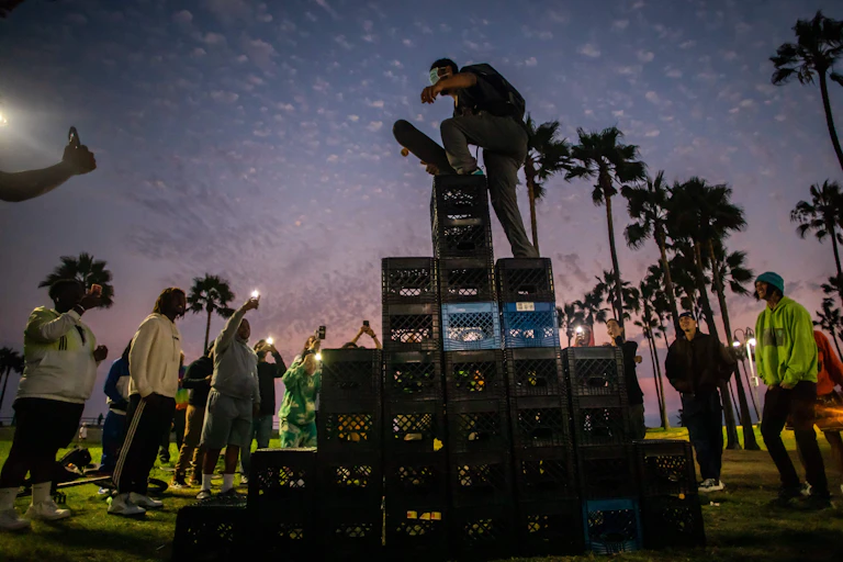Man with skateboard climbs up a stack of milk crates while people on ground film on their phones