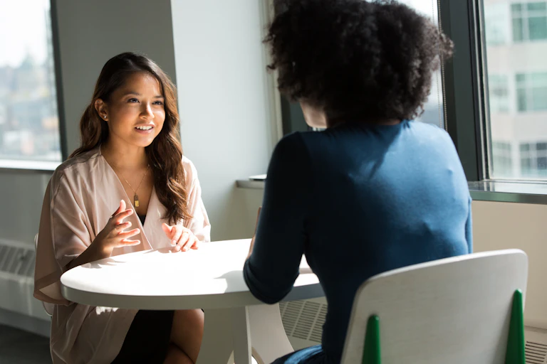 A young woman seated at a table talking to another woman with her back to the camera