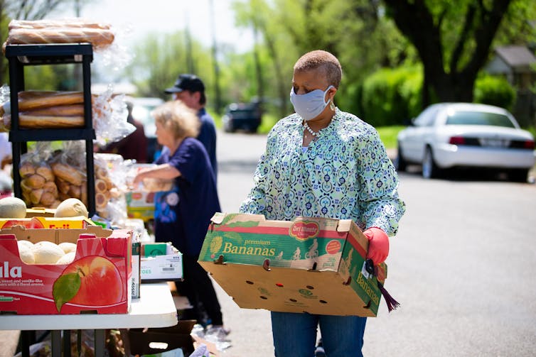 A woman with short hair wearing a mask carries a box of fresh fruit.