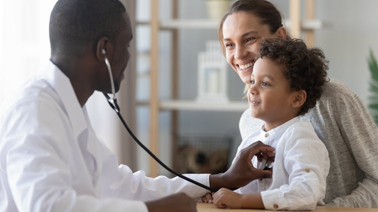 A woman with a child on her lap in a doctor's office, with the doctor holding a stethoscope to the child's chest