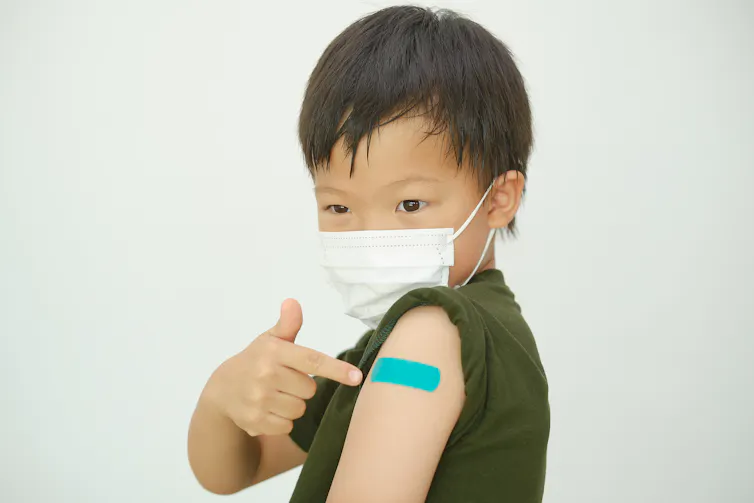 A child in a face mask pointing to a blue bandage on his arm