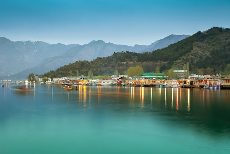 A town on the banks of a lake with light blue waters. Green mountains are seen in the background.