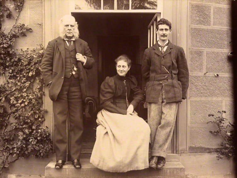 A black and white photo of three people in front of a house.