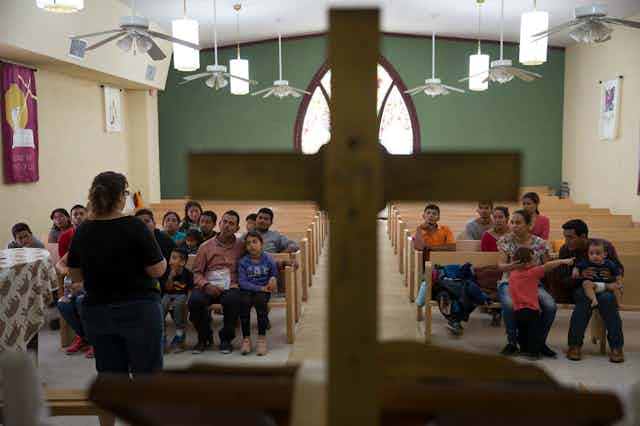 A woman standing at the front of a room with a cross in it speaks to a small crowd of people in pews.