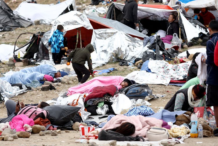 People sleep in the open air under blankets in a dry, dusty place.