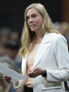 A woman with blonde hair wearing a white suit jacket speaking while standing.