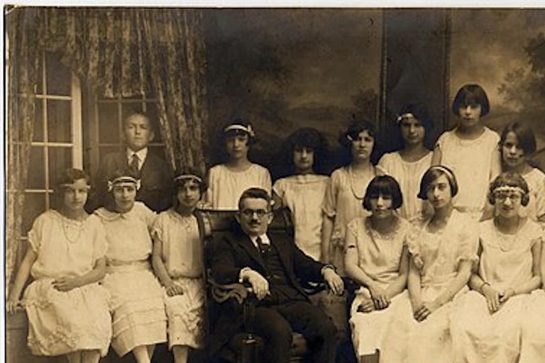 A group of young teens, mostly girls in white dresses, pose around a middle-aged man in a suit in a formal black and white portrait.