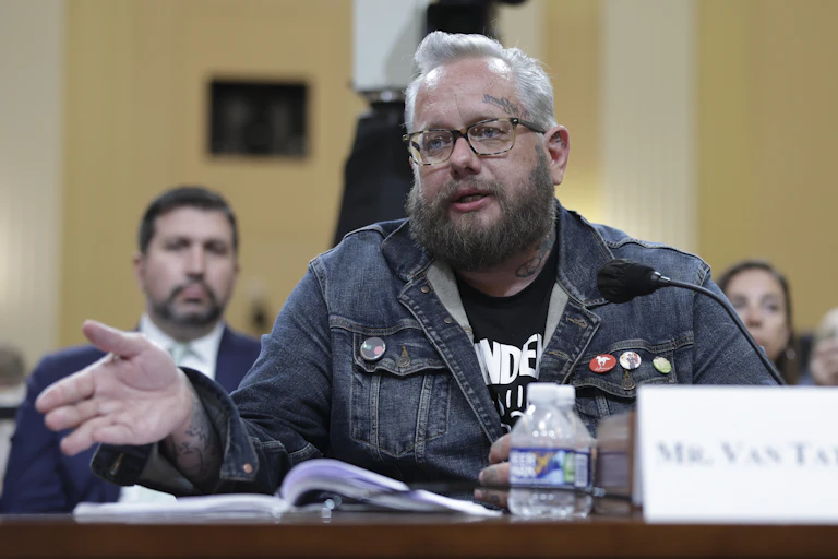 Man with glasses, a heavy beard and facial tattoos wearing informal clothing in a fancy setting.
