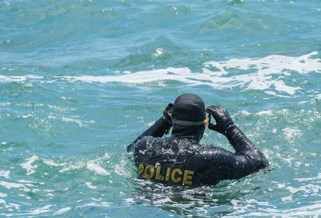 A diver in a wet suit that says police on the back standing in choppy sea water