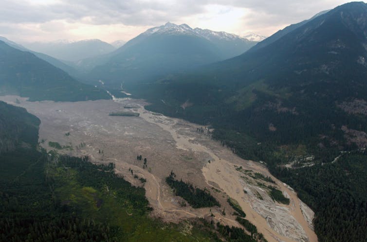 Smoke from wildfires hangs in the air as mud and debris fills a valley along a river