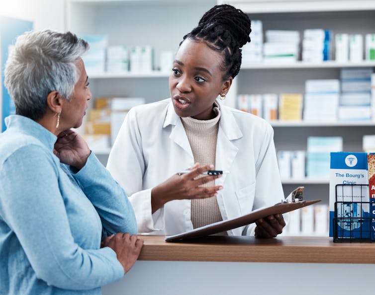 A young female pharmacist holding a clipboard and pen speaks with an older female customer.
