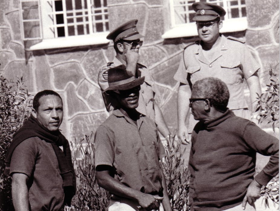 A black and white photograph of two prison guards standing supervising three men with gardening implements - a man on the left looking directly to camera.