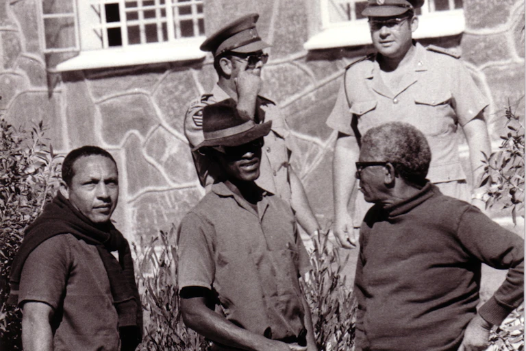 A black and white photograph of two prison guards standing supervising three men with gardening implements - a man on the left looking directly to camera.