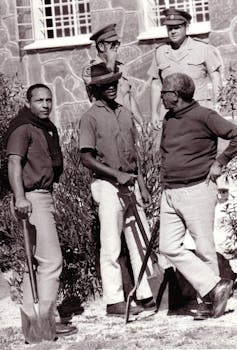 A black and white photograph of two prison guards standing supervising three men with gardening implements - a man on the left looking directly to camera.