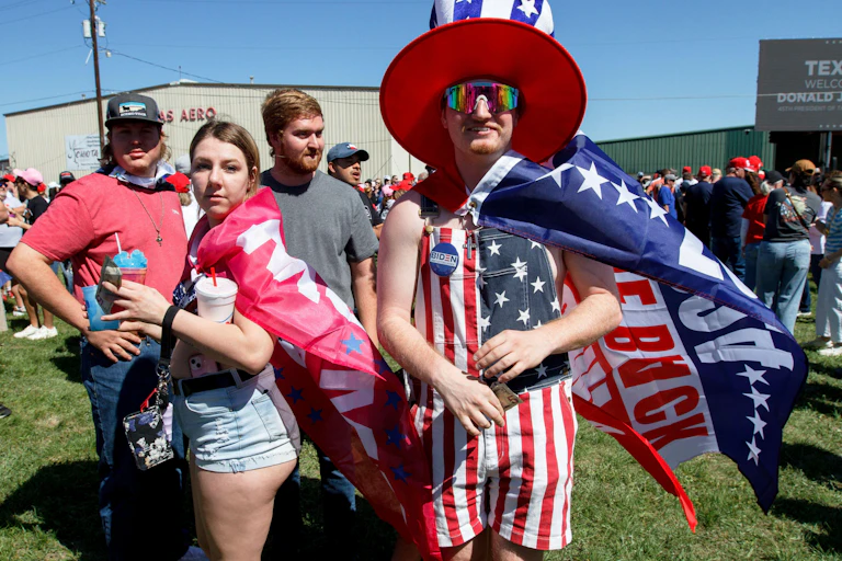 Two Donald Trump supporters wearing flags, one with a hat, looking towards the camera.