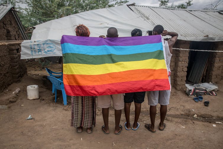 Four people stand, their backs to camera, holding a rainbow flag draped across their shoulders.