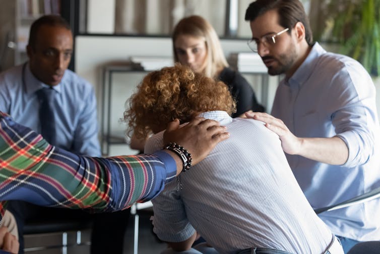 View of the back of a young woman sitting slumped at a table, while her colleagues pat her on the back in support