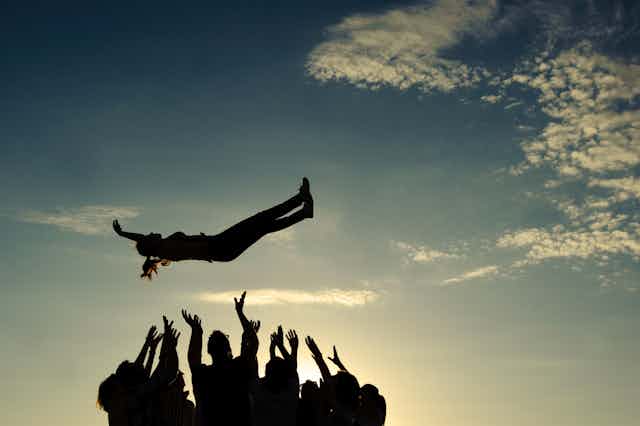 Silhouette of a group of people throwing a young woman in the air, with their arms outstretched to catch her